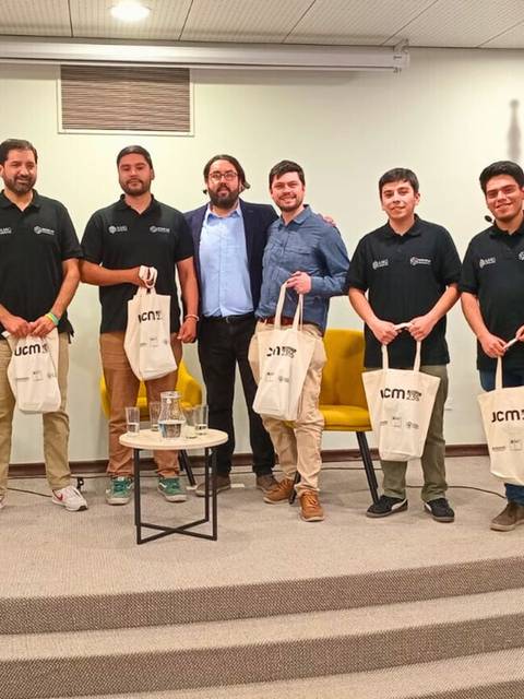 Un grupo de personas posando en un evento con mochilas, rodeados de banderas y un banner institucional.