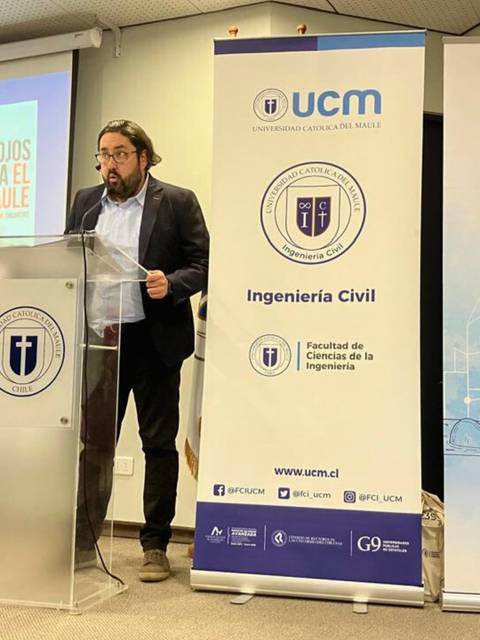 Un ponente discursando en un evento de la universidad con pantallas y banners visibles.