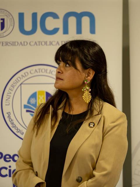 Una mujer con aretes grandes está mirando hacia un lado frente a una pared con logos de la universidad.
