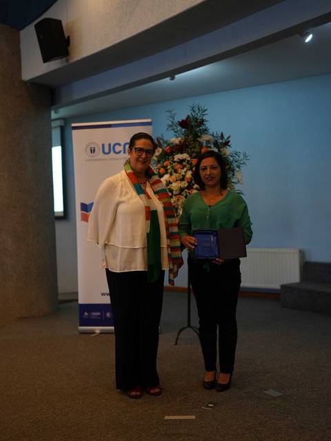 Dos mujeres posan sonriendo en un evento, una de ellas sostiene un reconocimiento en sus manos.