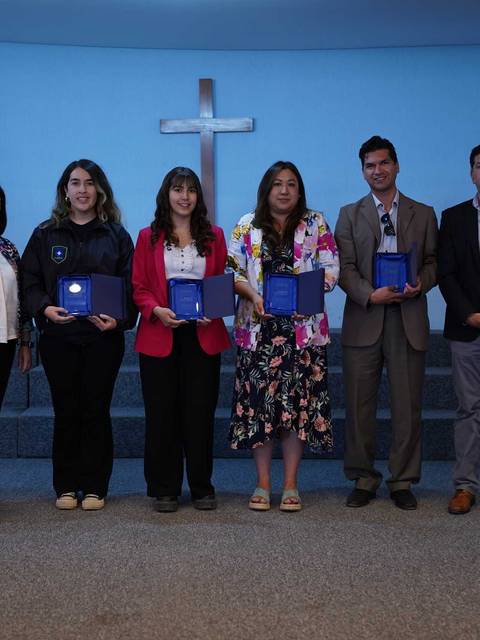 Un grupo de seis personas posan en un escenario con diplomas en las manos frente a una cruz en la pared.