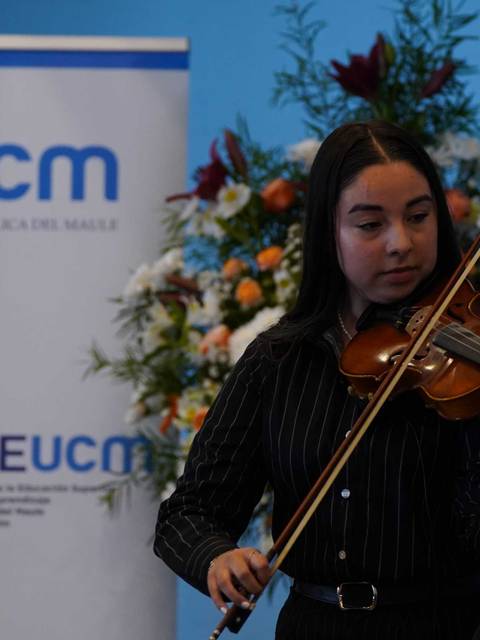 Una joven tocando el violín frente a una pancarta de la Universidad Católica del Maule con un arreglo floral al fondo.