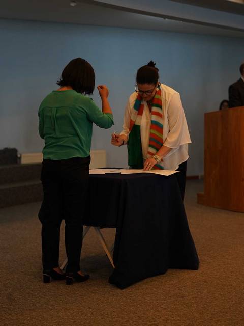 Dos personas se encuentran en un ambiente formal, colaborando en un documento frente a un estrado y una bandera.