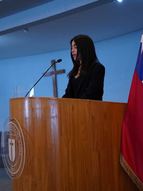 Una mujer se encuentra de pie frente a un podio en un evento formal, con la bandera de Chile al lado.