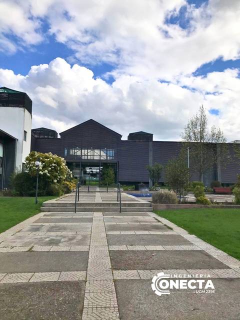 La imagen muestra un paseo con escaleras que conduce a un edificio moderno rodeado de jardines y nubes en el cielo.