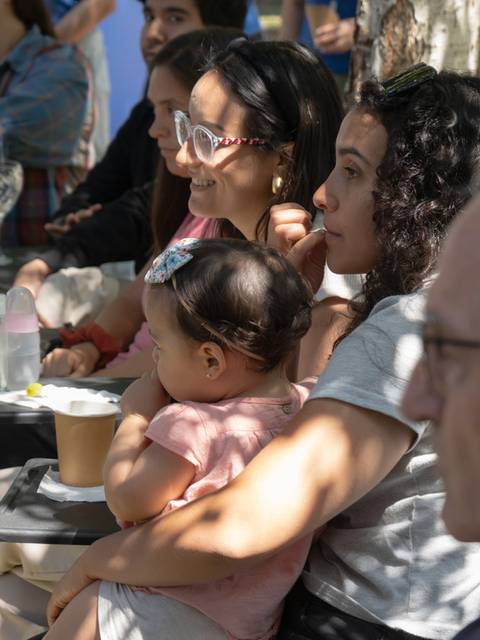 Un grupo de personas sentadas al aire libre, algunas con bebés, escuchando atentamente.