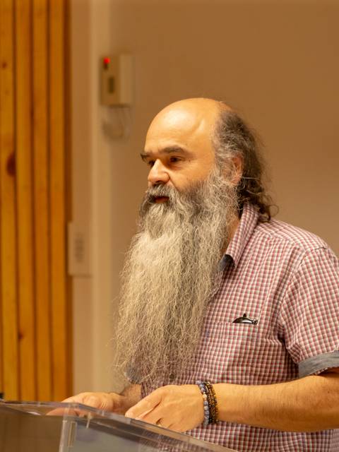 Un hombre con barba larga y canosa habla frente a un micrófono en un evento.