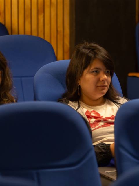 Dos mujeres sentadas en un auditorio con sillas azules.