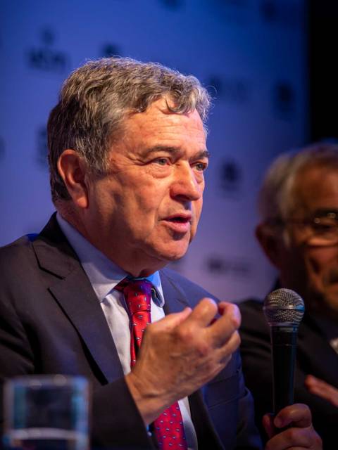 Un hombre con cabello gris habla durante un panel, mientras otro hombre lo escucha atentamente.
