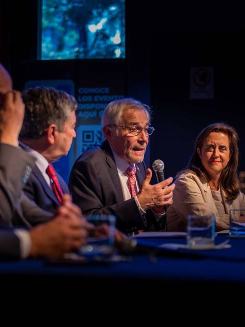 Un grupo de personas sentadas en una mesa durante un evento, con un hombre hablando y el resto escuchando atentamente.