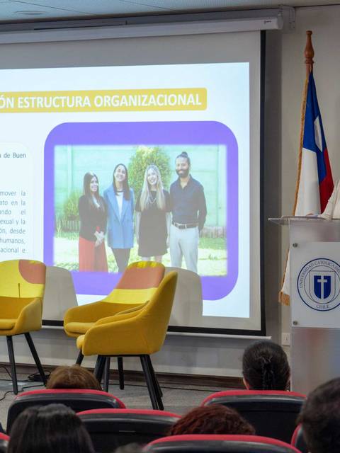 Una presentadora en un evento está hablando frente a una pantalla que muestra información sobre la actualización de una estructura organizacional.