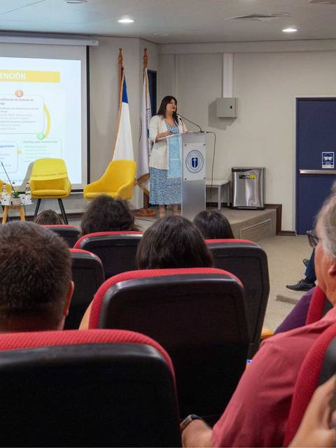 Una presentadora expone frente a una audiencia en un auditorio con sillas rojas y amarillas.
