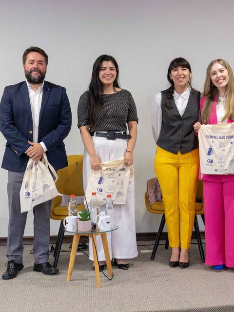 Cuatro personas posan juntas en un evento, sosteniendo bolsas, con sillas amarillas de fondo.