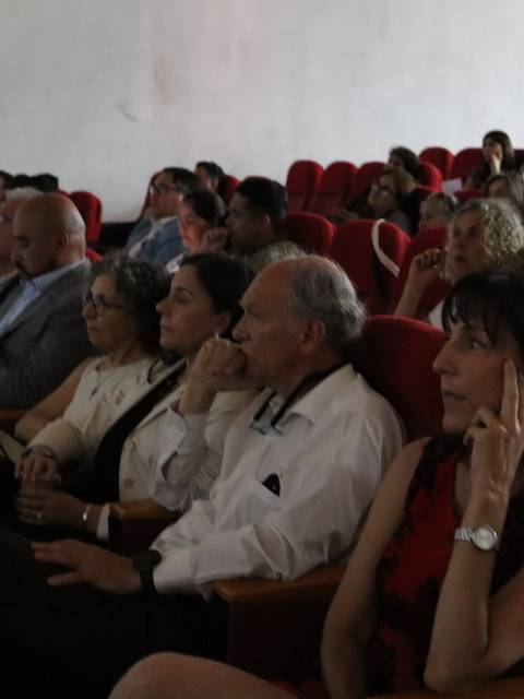 Una audiencia presta atención en un evento en un salón con asientos rojos.