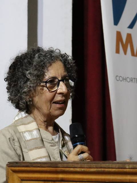 Una mujer con cabello rizado y gafas está hablando en un evento mientras sostiene un micrófono.