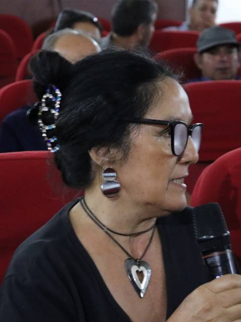 Una mujer con gafas y peinado característico sostiene un micrófono mientras charla en un auditorio lleno de gente.