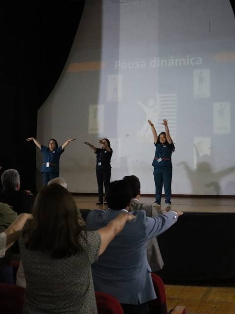 Un grupo de personas está participando en una pausa dinámica en un evento, mientras siguen las instrucciones de tres presentadoras en el escenario.