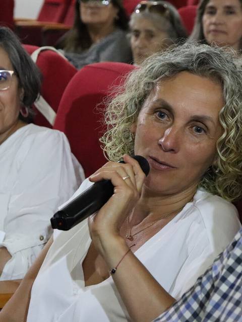 Mujer hablando con un micrófono en una charla, rodeada de asistentes en un auditorio.
