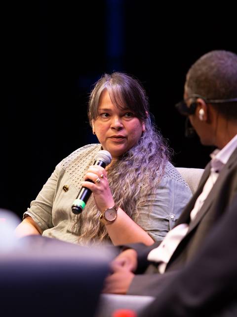 Una mujer habla en un panel utilizando un micrófono mientras escucha a un colega.