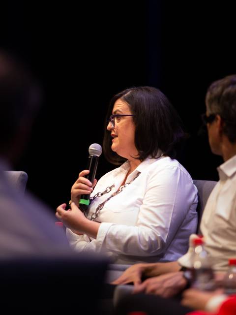 Una mujer habla en un panel durante un evento, sosteniendo un micrófono mientras otros participantes la escuchan.