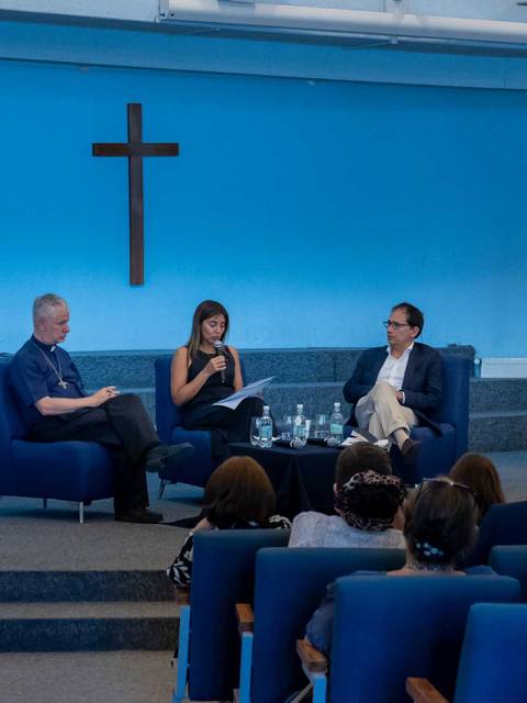 Una conferencia en un auditorio con un crucifijo en la pared, donde tres personas están sentadas y el público está atento.