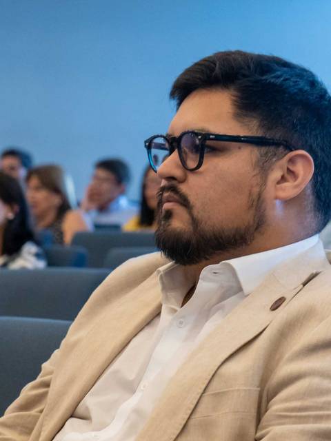 Un hombre con gafas y un traje claro escucha atentamente en un evento con audiencia.