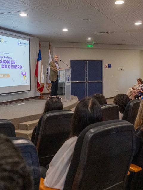 Una conferencia sobre la Comisión de Equidad de Género con un orador frente a una audiencia en un auditorio.