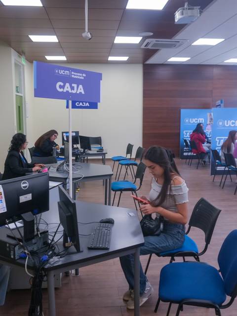 Un aula moderna con varias mesas de trabajo y personas interaccionando con computadoras y teléfonos móviles.