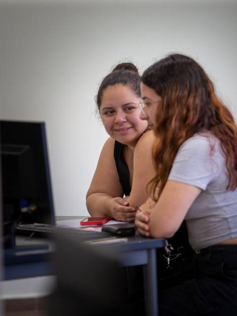 Dos jóvenes conversando y sonriendo en un entorno académico con computadoras.