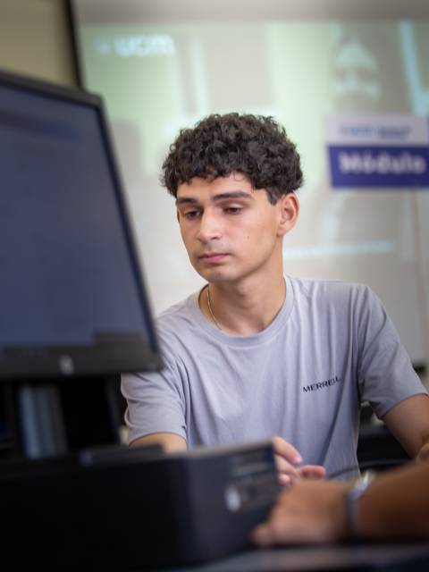 Un joven está sentado frente a un computador en un aula con otros estudiantes en el fondo.