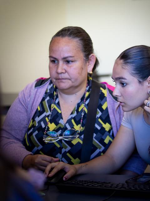 Dos mujeres están concentradas frente a un ordenador, una de ellas parece estar ayudando a la otra.