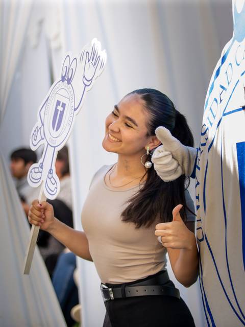 Una joven sonríe mientras sostiene un cartel y hace un gesto positivo en un evento.