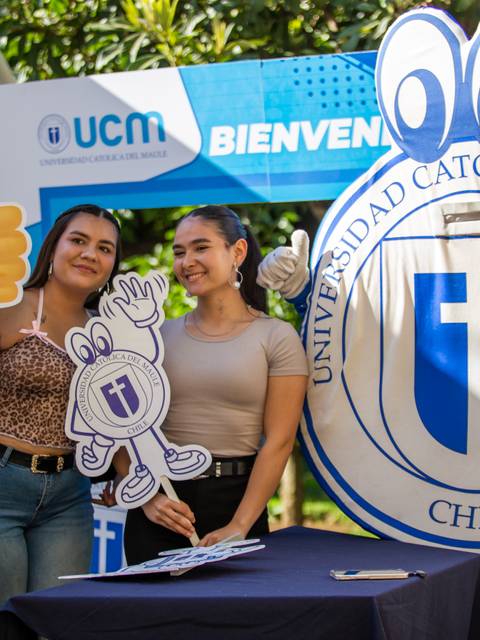 Dos estudiantes posan juntas con carteles y un fondo de la Universidad Católica del Maule.