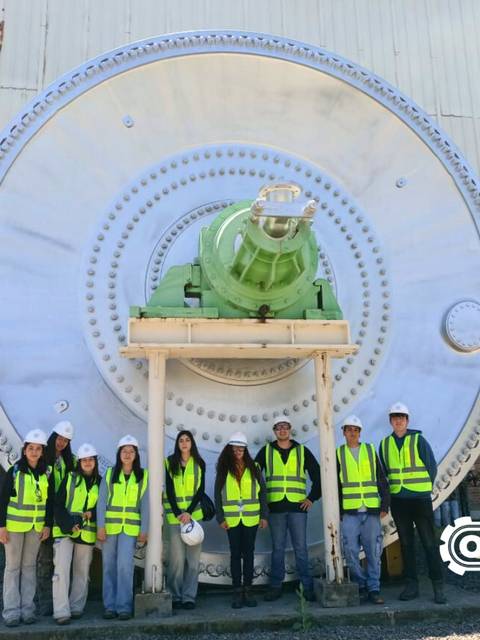 Grupo de personas con chalecos amarillos posando frente a una gran rueda industrial.