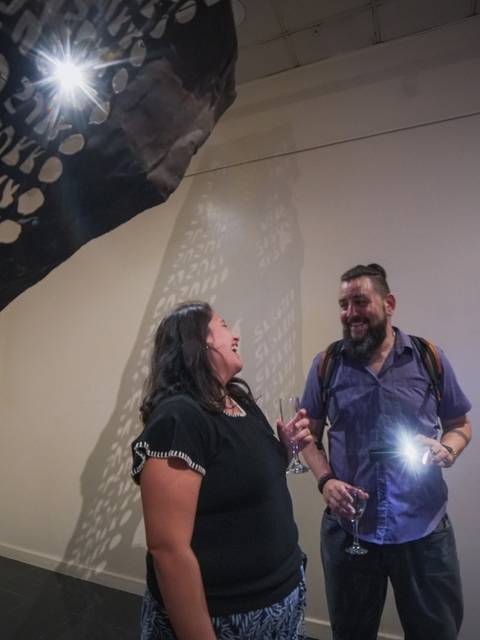 En la imagen, dos personas están explorando una instalación artística que proyecta sombras de textos en una pared.