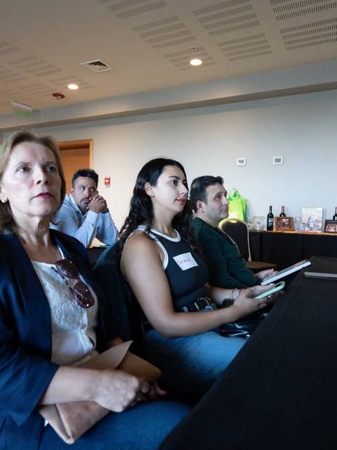 Una reunión con público atento en una sala con mesas y bebidas al fondo.