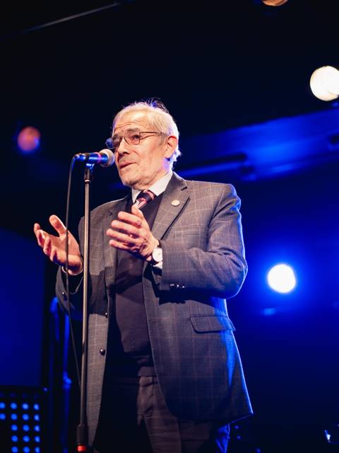 Un hombre mayor canta con pasión en un escenario iluminado.