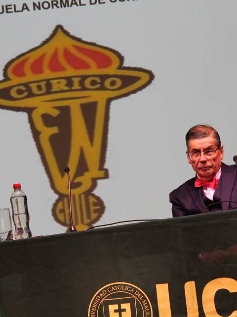 Un hombre mayor sentado en una mesa con un logo de la Escuela Normal de Curicó de fondo.