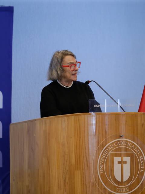Una mujer con gafas rojas habla desde un podio en un evento en la Universidad Católica de Chile.