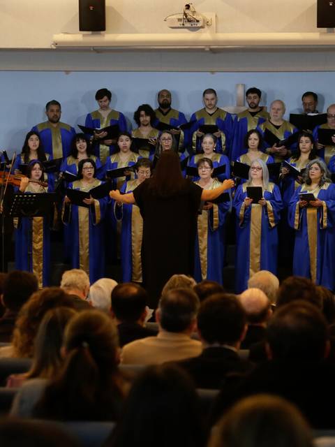 Un grupo de personas con túnicas azules está cantando en un escenario frente a un público atento.