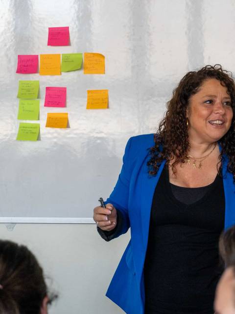 Una mujer con cabello rizado y chaqueta azul está hablando frente a un grupo de personas durante una dinámica en una sala de clases.