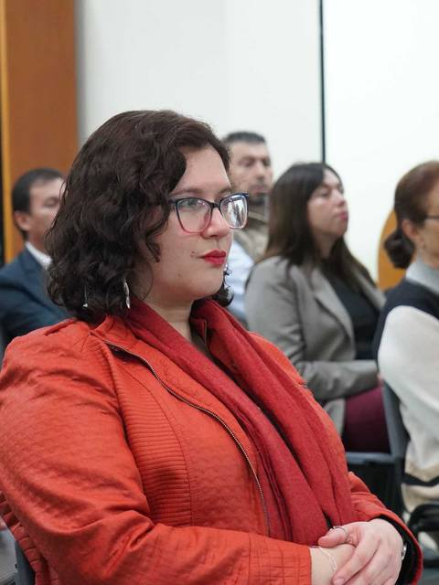 En una reunión, la gente escucha atentamente mientras una mujer con un abrigo naranja se destaca entre los asistentes.