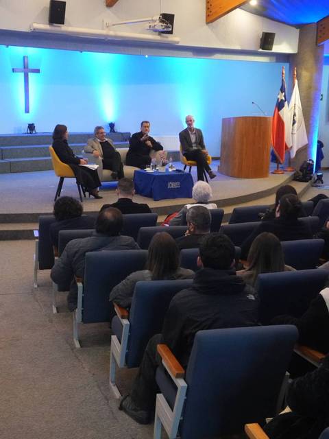Un panel de discusión en una sala de conferencias en Chile, con un público atento y una cruz iluminada de fondo.