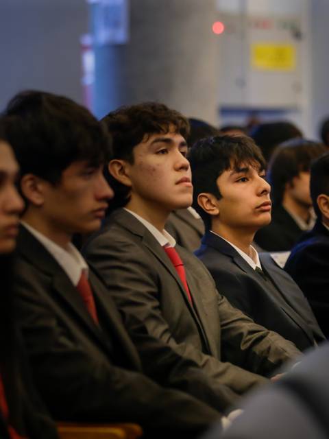 Un grupo de estudiantes sentados en un evento formal, vistiendo trajes y con atención en el escenario.