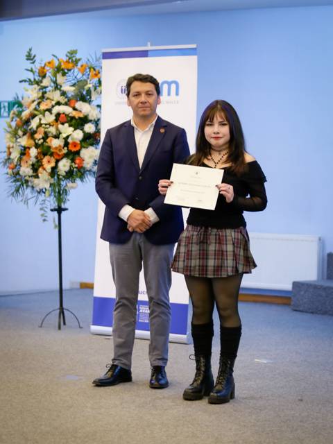 Una joven sonriente sostiene un certificado junto a un hombre en un evento formal.