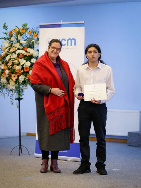 Un joven recibe un reconocimiento en una ceremonia formal, acompañado por una mujer con una chaqueta roja.