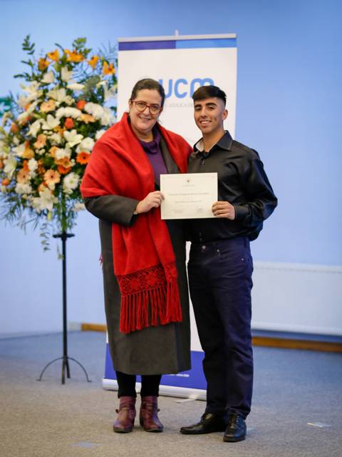 Una ceremonia de graduación donde un joven recibe un certificado de manos de una mujer con un chal rojo.