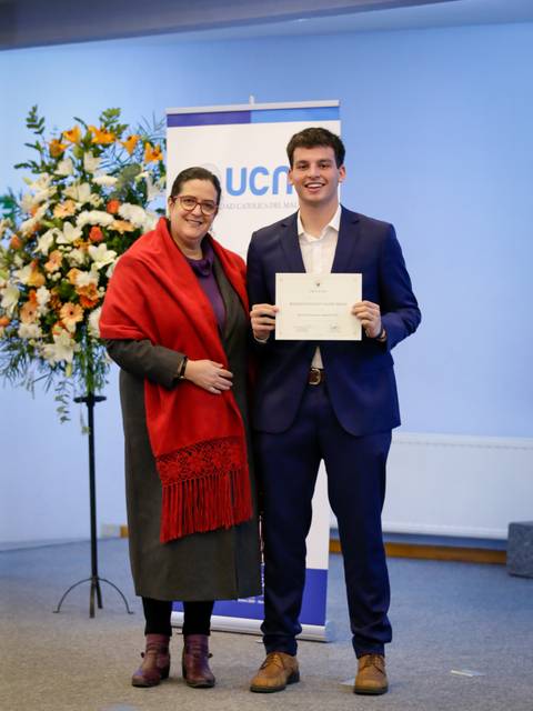 Un joven sonriente recibe un certificado de manos de una mujer vestida con un chal rojo en un evento oficial.