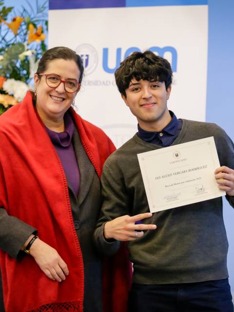 El cabro chico está recibiendo un diploma y sonríe feliz junto a una profe en un evento.