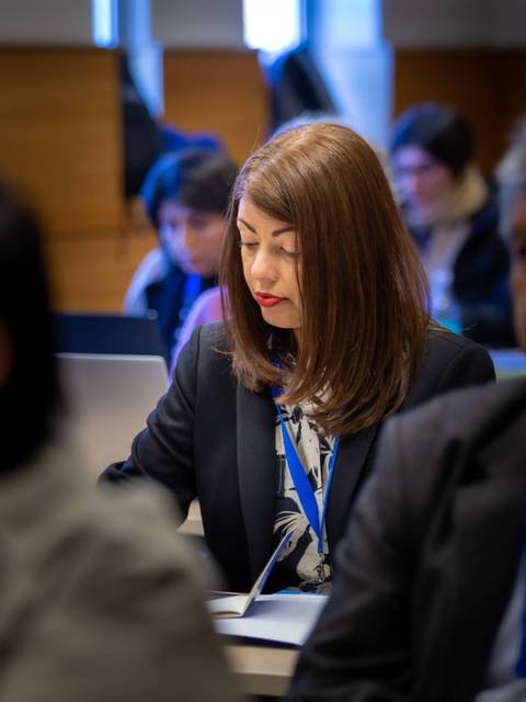 Una mujer con cabello largo y castaño está tomando apuntes en una reunión mientras otros asistentes la observan.